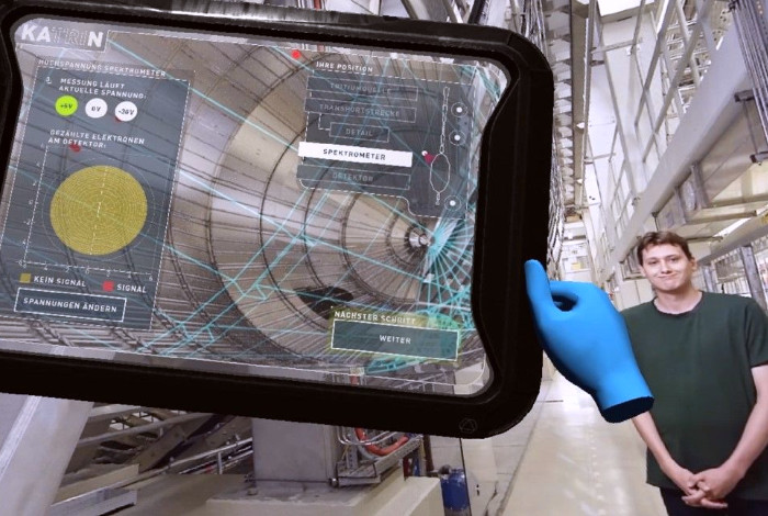 Einblick in die VR-Anwendung: Der Physiker Dr. Manuel Klein erklärt neben dem Hauptspektrometer-Tank das KATRIN-Experiment. Mit dem virtuellen Tablet können die Benutzerinnen und Benutzer der VR-Anwendung das Experiment steuern. (Quelle: Screenshot aus de